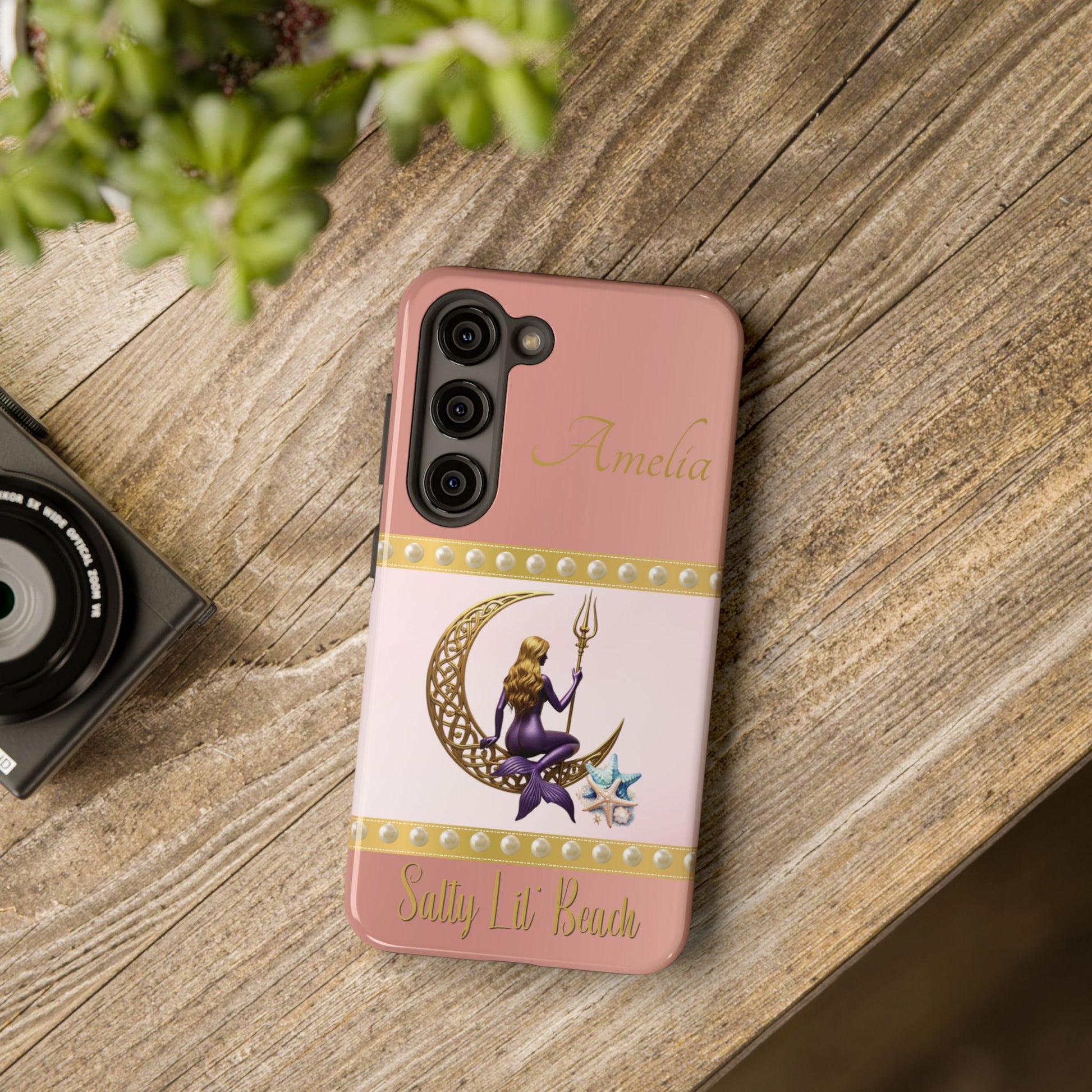 Phone case with mermaid design on a wooden surface next to a camera and plant