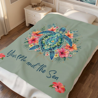 A sea turtle and floral design printed on a sage green bedspread or blanket, styled on a bed in a bedroom setting. The design features a blue and green sea turtle surrounded by pink, orange, and yellow tropical flowers on a sage green background, with the text 'You Me and the Sea' in blue script.