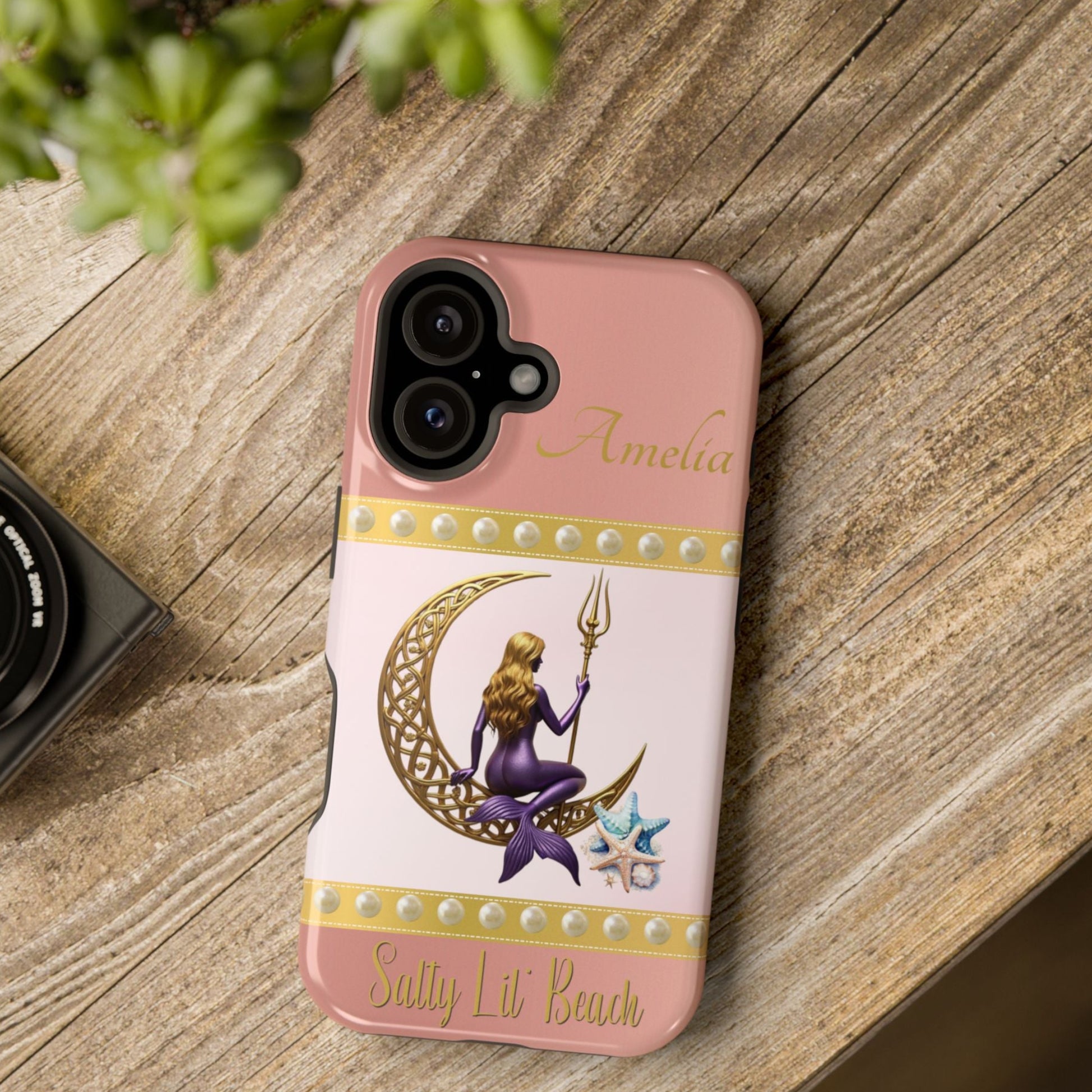 Phone case with a design of a mermaid and crescent moon on a pink background, placed on a wooden surface.