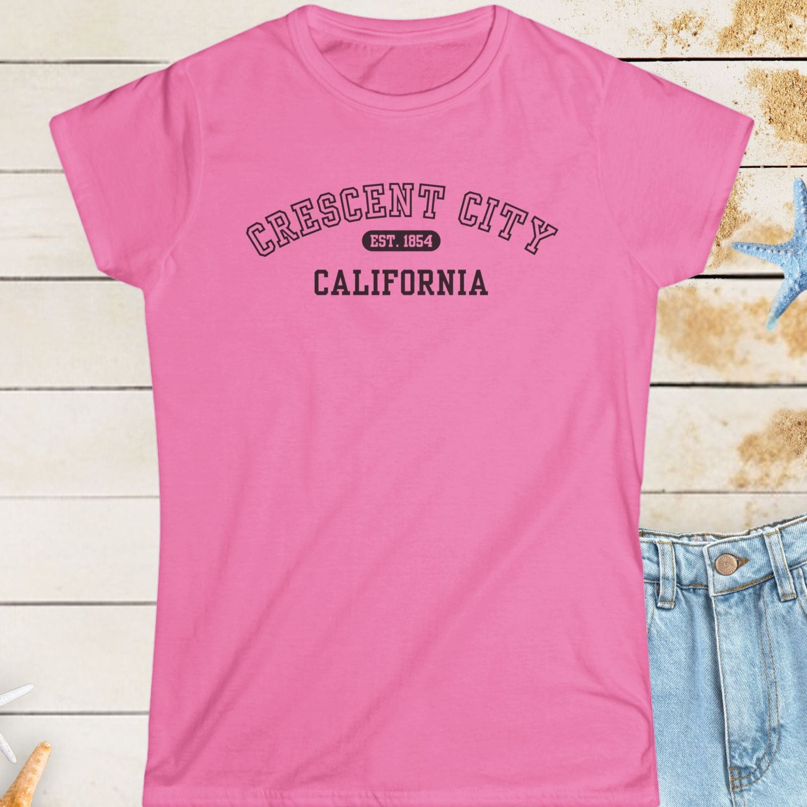Pink t-shirt with 'Crescent City, California' text on a wooden surface with starfish.