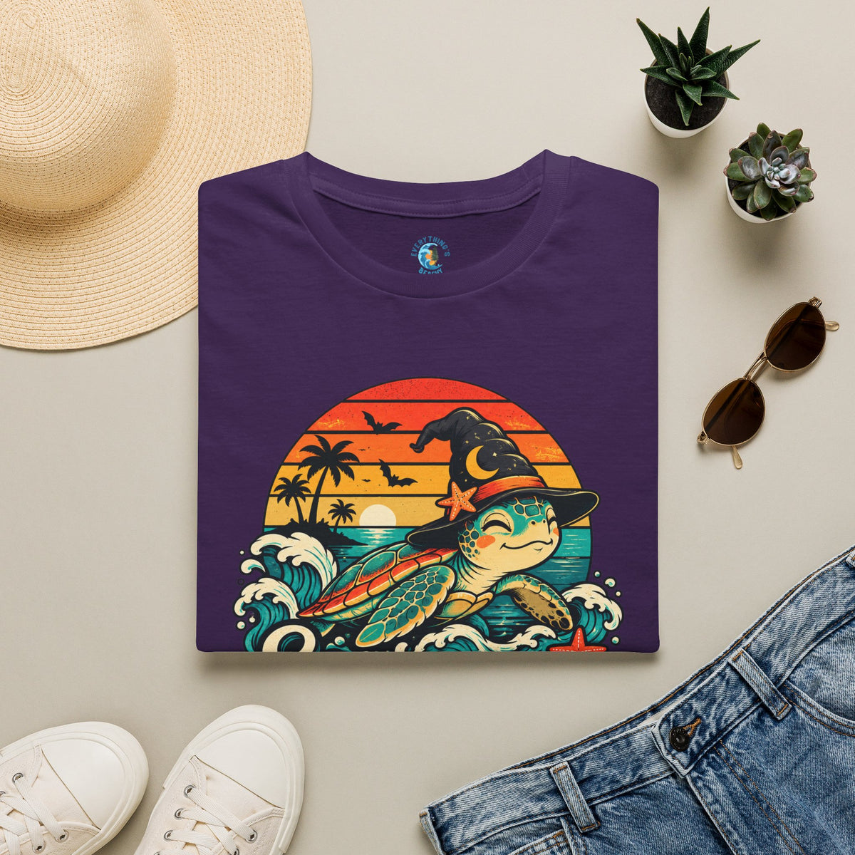 Folded Purple unisex t-shirt with “Seas the Magic” text, a sea turtle wearing a witch hat, ocean waves, sunset, palm trees, bats, and starfish.