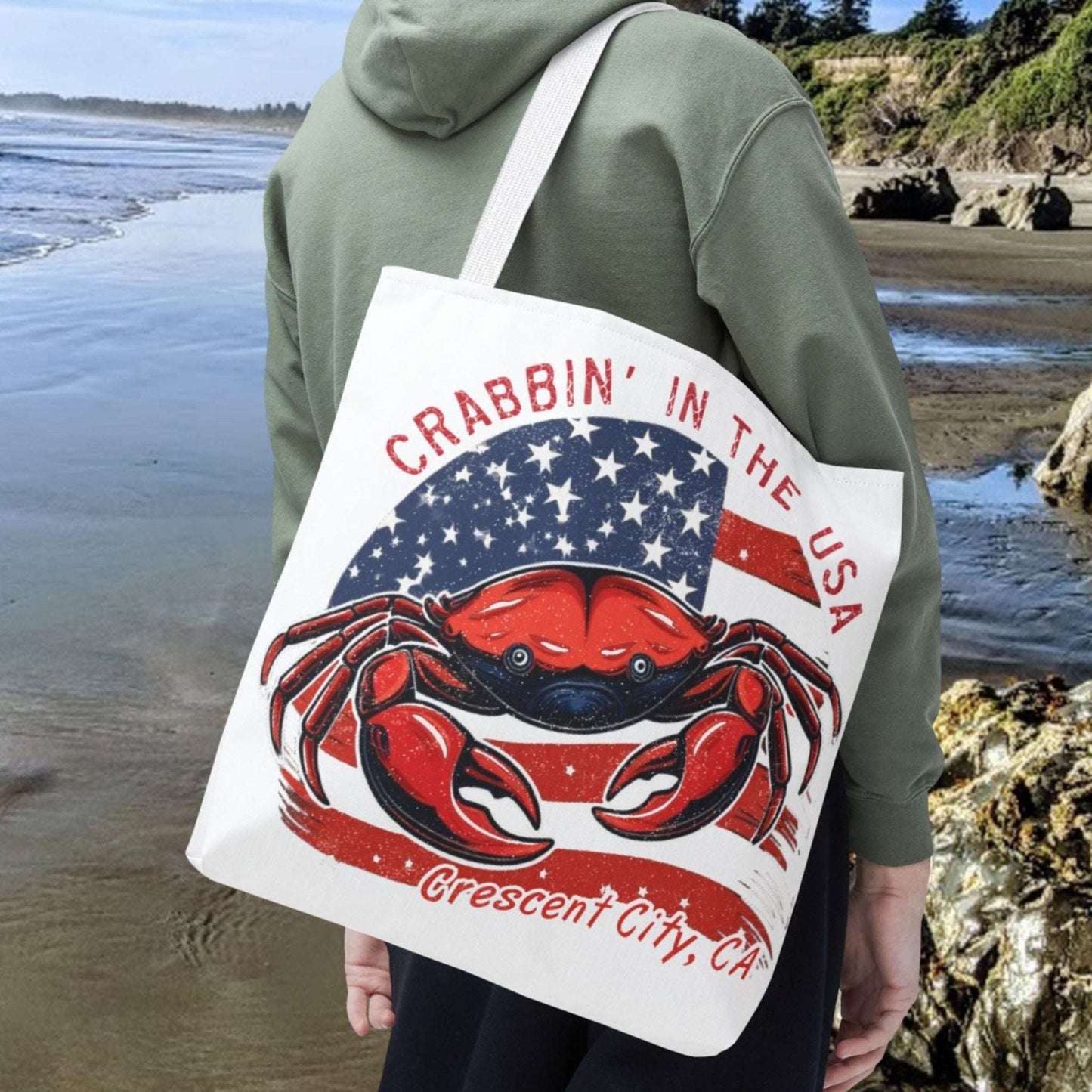 Man on a beach holding a tote bag with a red crab design and text Crabbin' in the USA Crescent City, CA.