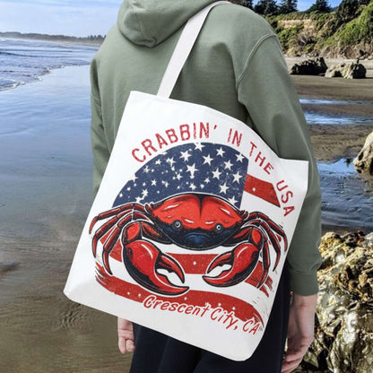 Man on a beach holding a tote bag with a red crab design and text Crabbin' in the USA Crescent City, CA.