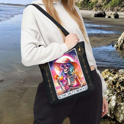 Person holding a tote bag with a colorful skeleton woman holding a cocktail with a colorful beach sunset on a tarot card design with text The Salty Beach.