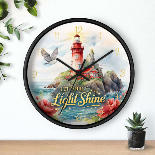 Decorative wall clock with lighthouse design and 'Let Our Light Shine' text, placed on a surface with plants.