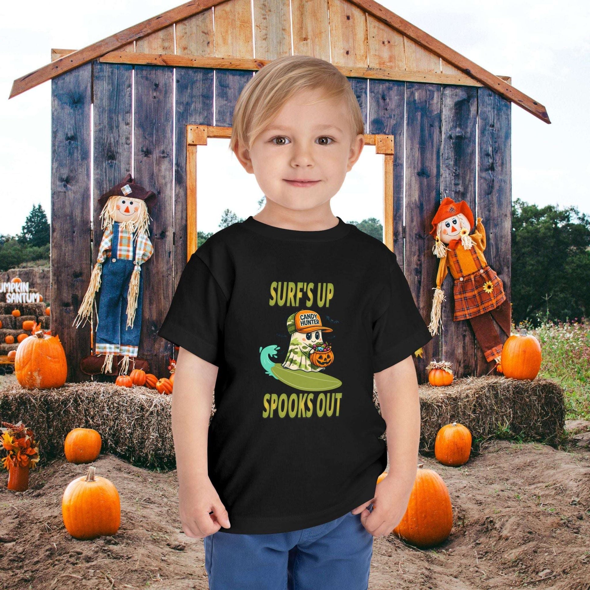 Child wearing a black toddler t-shirt with a cartoon ghost child on a surfboard, holding a pumpkin candy bucket, wearing an orange hat labeled “Candy Hunter,” with text that reads “Surf’s Up Spooks Out.”