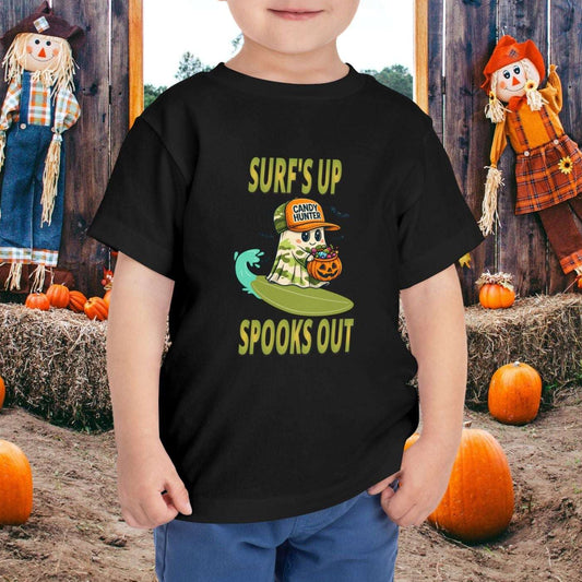 Child wearing a black toddler t-shirt with a cartoon ghost child on a surfboard, holding a pumpkin candy bucket, wearing an orange hat labeled “Candy Hunter,” with text that reads “Surf’s Up Spooks Out.”