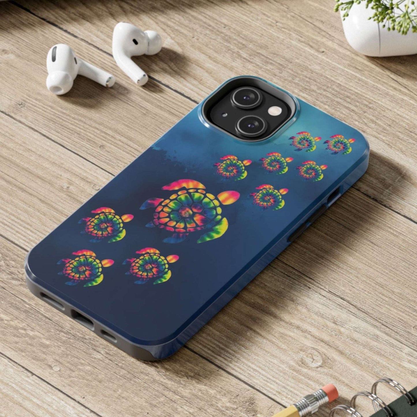 Blue phone case with colorful turtle design on a wooden desk with laptop, earbuds, and notebook.