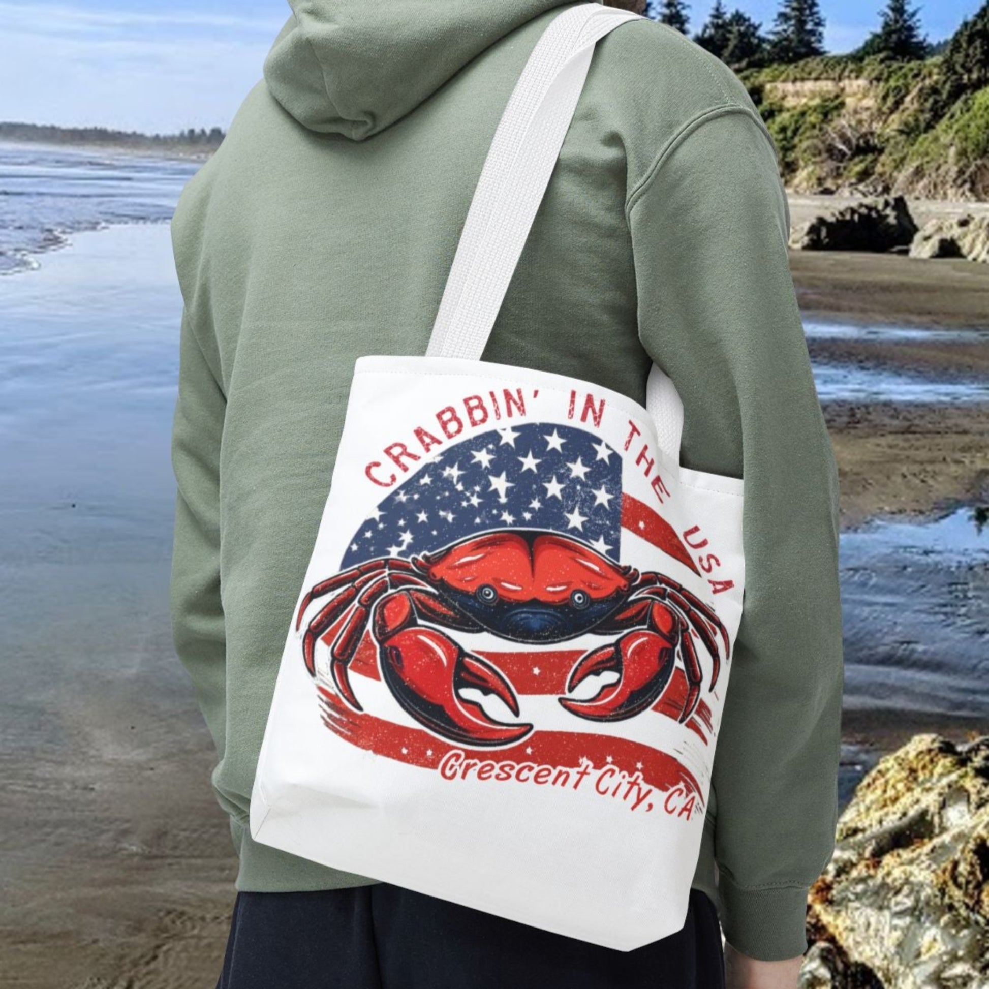 Person on a beach holding a tote bag with a crab design and text.