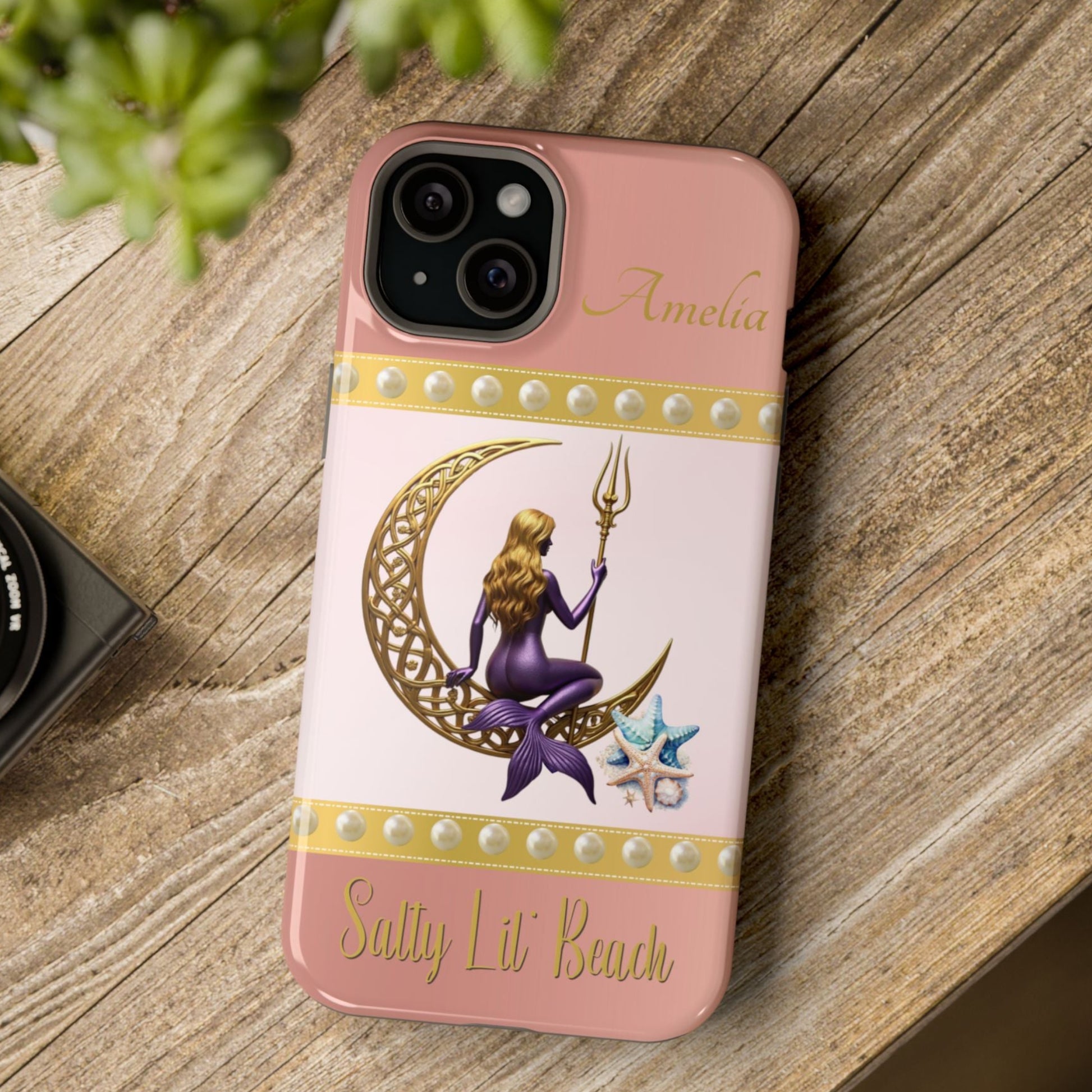 Phone case with a design of a mermaid and crescent moon on a pink background, placed on a wooden surface.