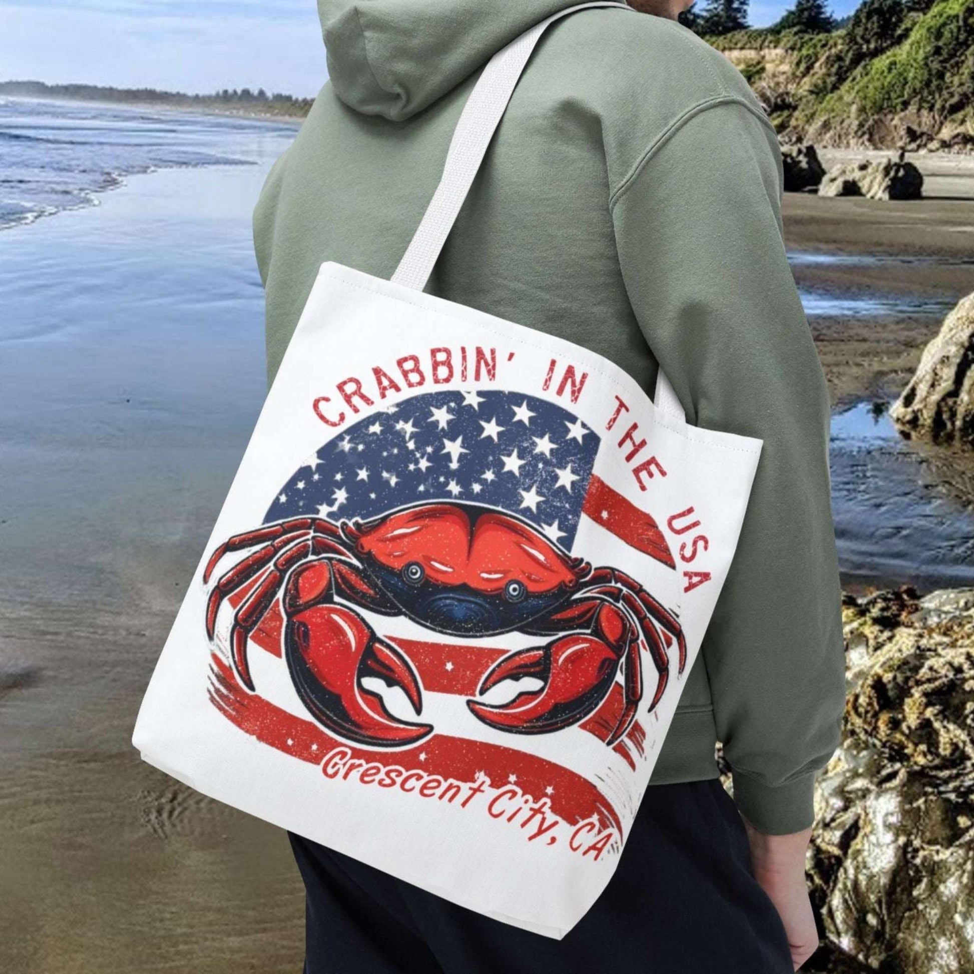 Person on a beach holding a tote bag with a red crab design and text Crabbin' in the USA Crescent City, CA.