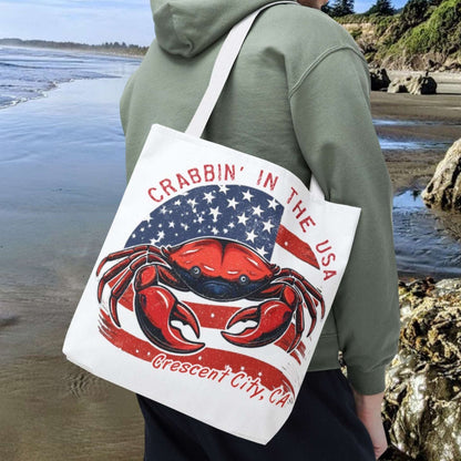 Person on a beach holding a tote bag with a red crab design and text Crabbin' in the USA Crescent City, CA.