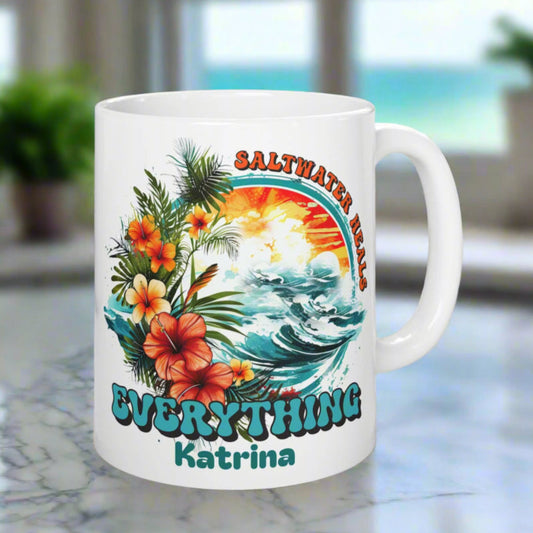 White mug with tropical design and text on a coaster, placed on a surface with a plant and container in the background.
