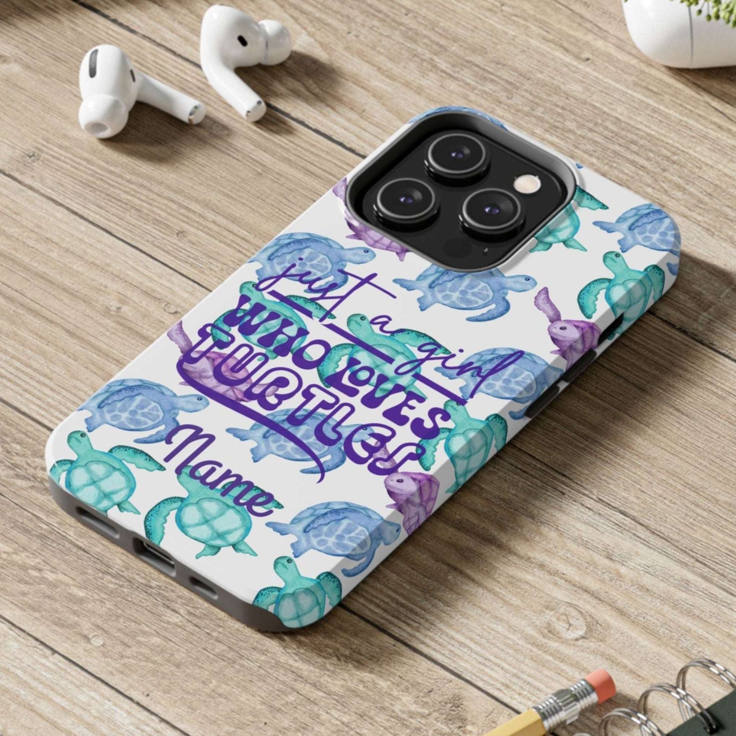 Phone case with sea turtle design and text on a wooden desk with laptop, earbuds, and notebook.