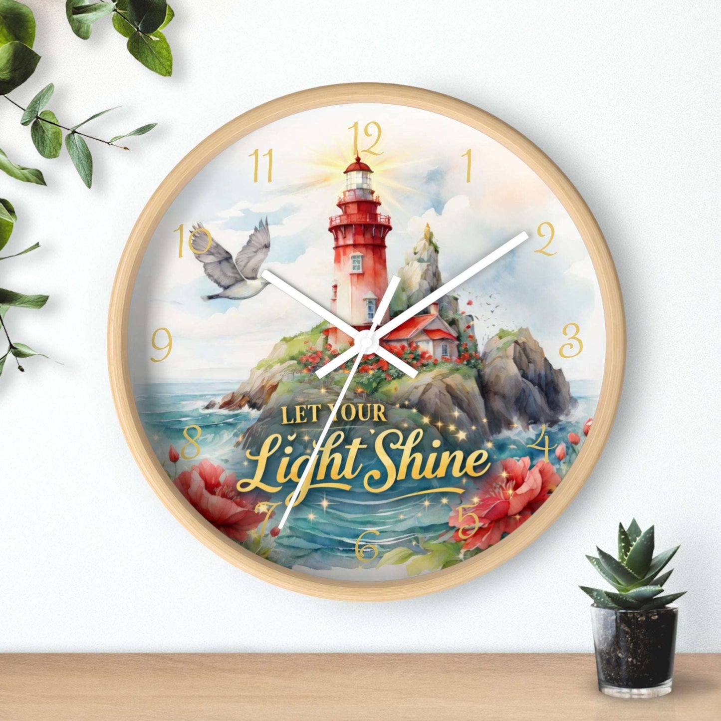 Decorative wall clock with a lighthouse design and 'Let Your Light Shine' text, surrounded by plants on a wooden surface.