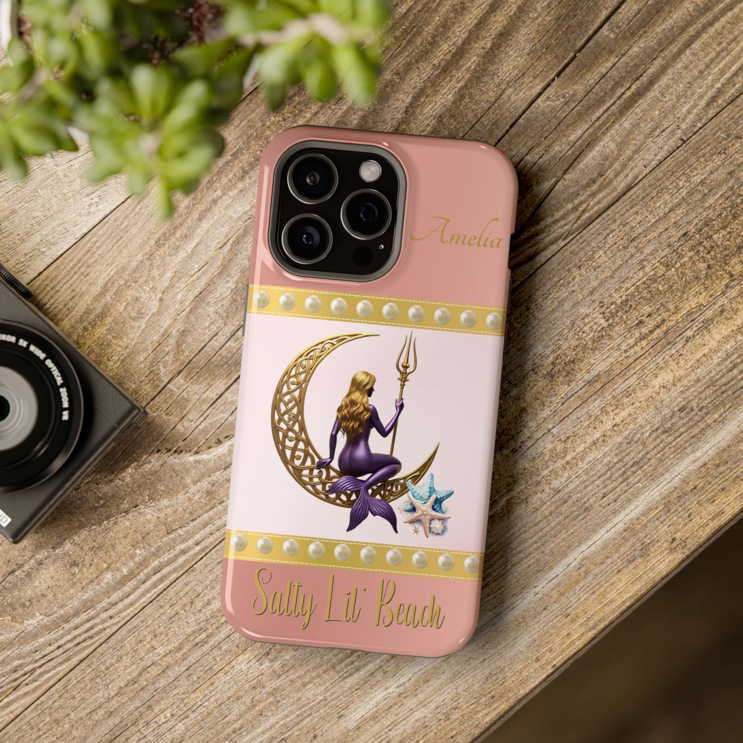 Phone case with a design of a mermaid and crescent moon on a pink background, placed on a wooden surface.