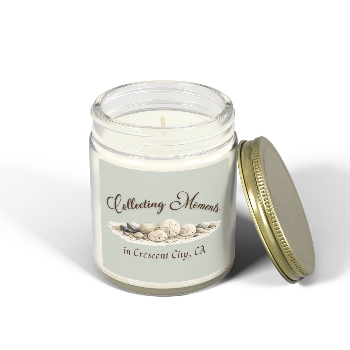 A glass jar candle with a gold metal lid sits on a wooden surface, surrounded by eucalyptus leaves. The candle has a sage green label that reads 'Collecting Moments in Crescent City, CA' with an illustration of seashells.