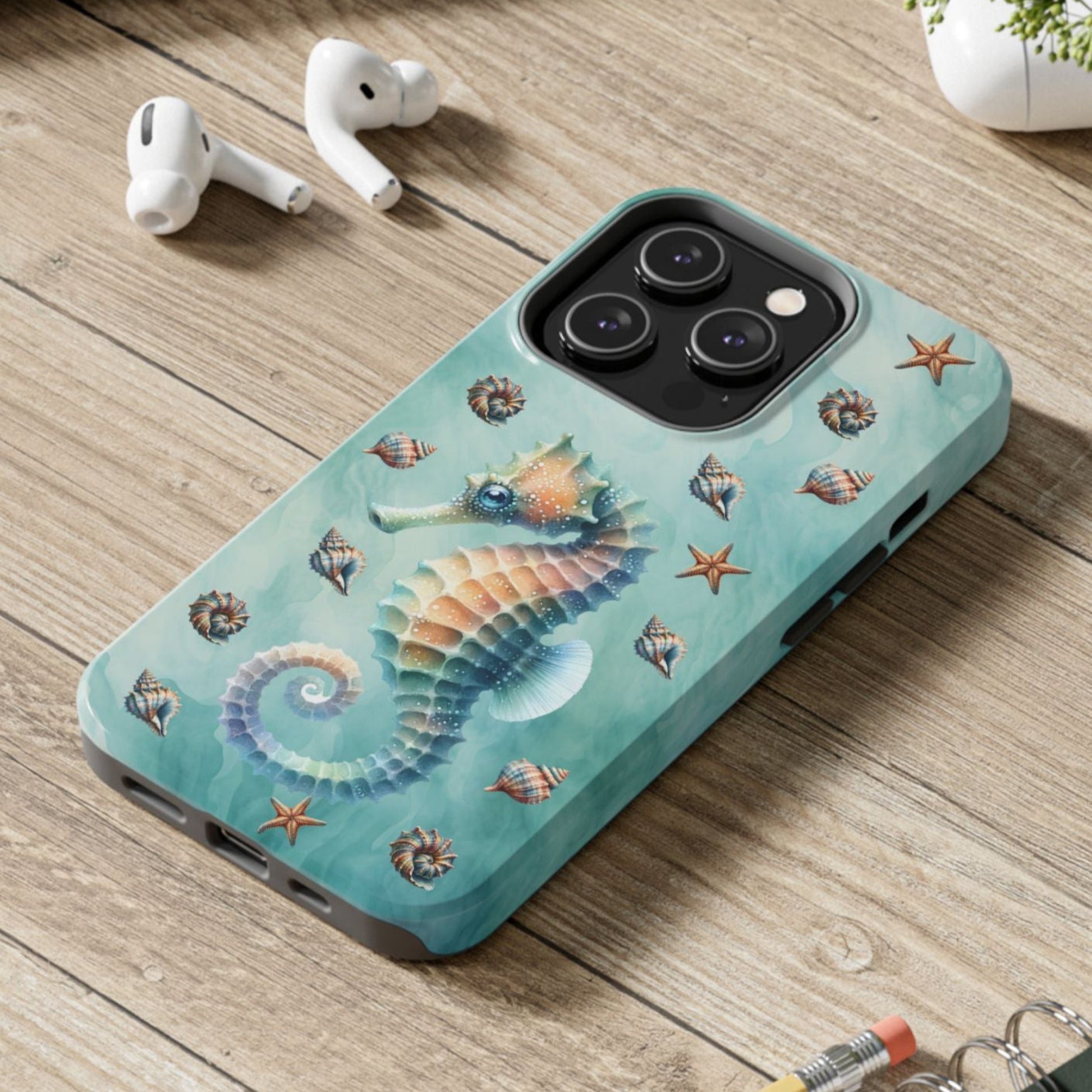 Phone case with seahorse design on a wooden surface with laptop, earbuds, and notebook.