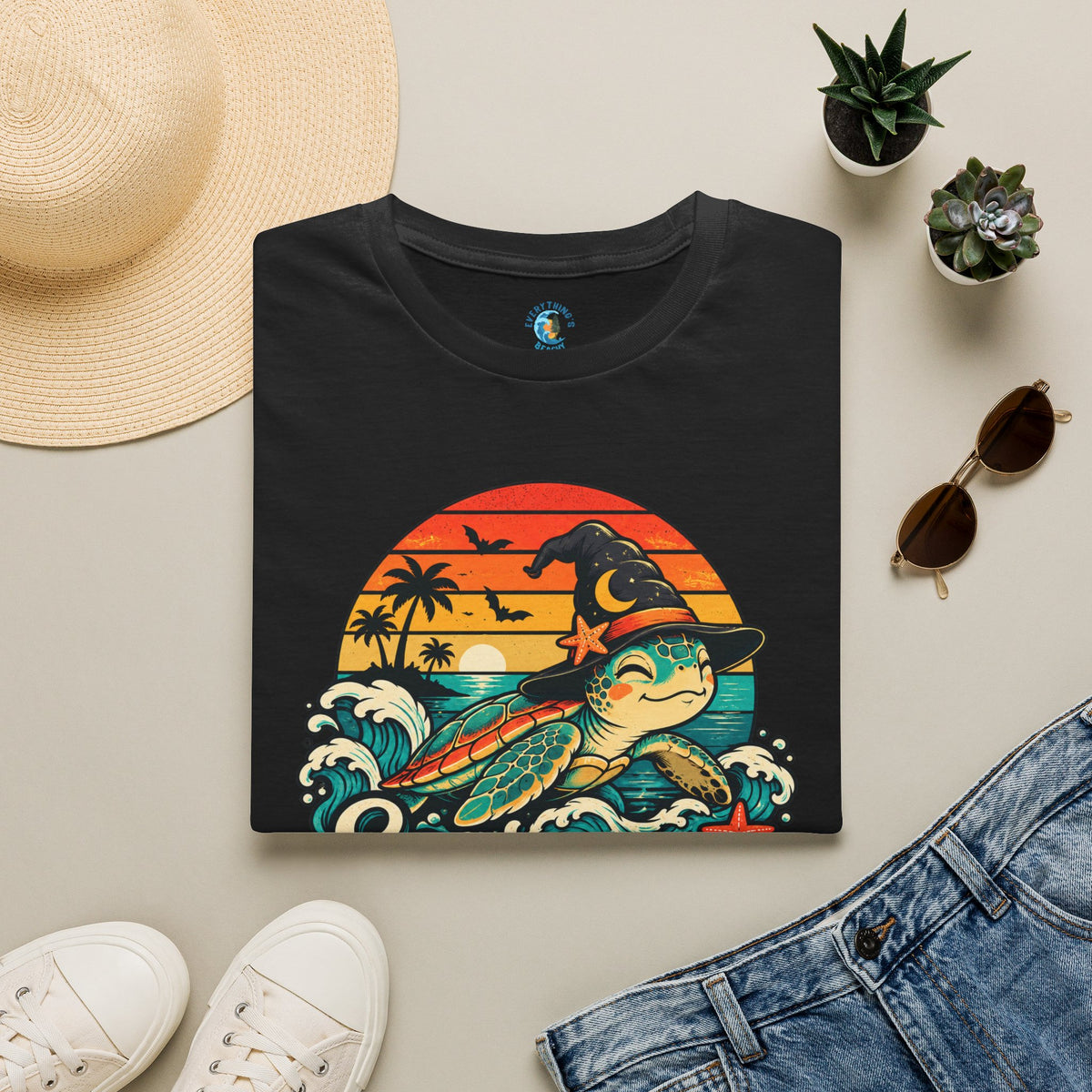 Folded Black unisex t-shirt with “Seas the Magic” text, a sea turtle wearing a witch hat, ocean waves, sunset, palm trees, bats, and starfish.