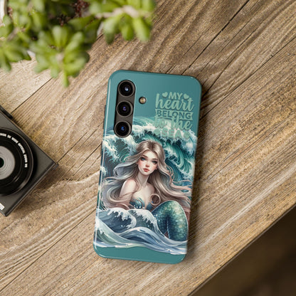 Phone Case with Flowy mermaid with ocean wave behind and text My Heart Belongs to the Sea laying on desk.