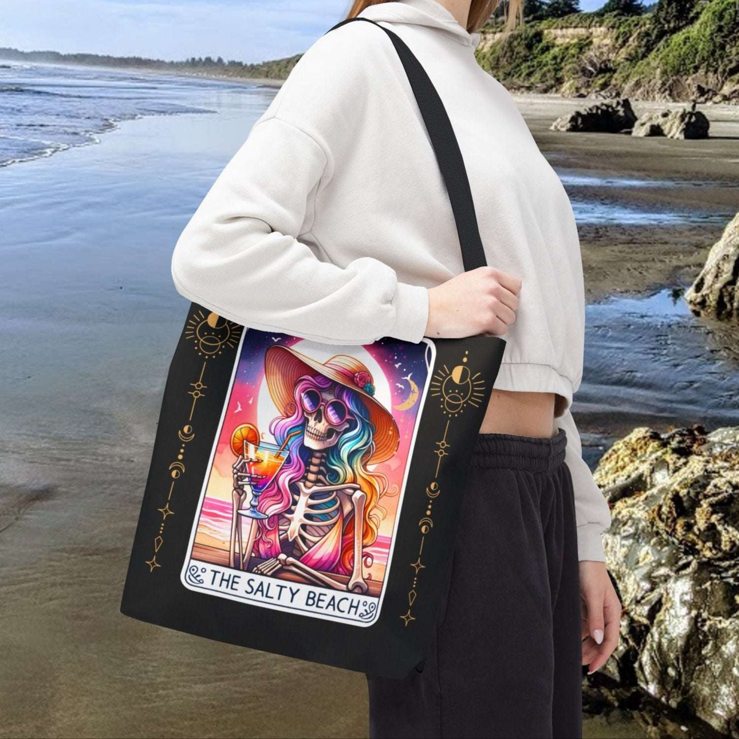 Person holding a tote bag with a colorful skeleton woman holding a cocktail with a colorful beach sunset on a tarot card design with text The Salty Beach.