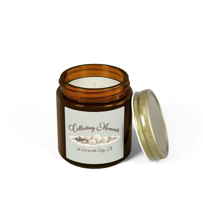 An amber glass jar candle with a gold metal lid sits on a wooden surface, surrounded by eucalyptus leaves. The candle has a sage green label that reads 'Collecting Moments in Crescent City, CA' with an illustration of seashells.