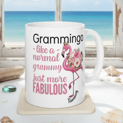 Ceramic coffee mug featuring a flamingo adorned with flowers and the text “Grammingo like a normal Grammy just more fabulous,” available with white, pink, or black handle options.