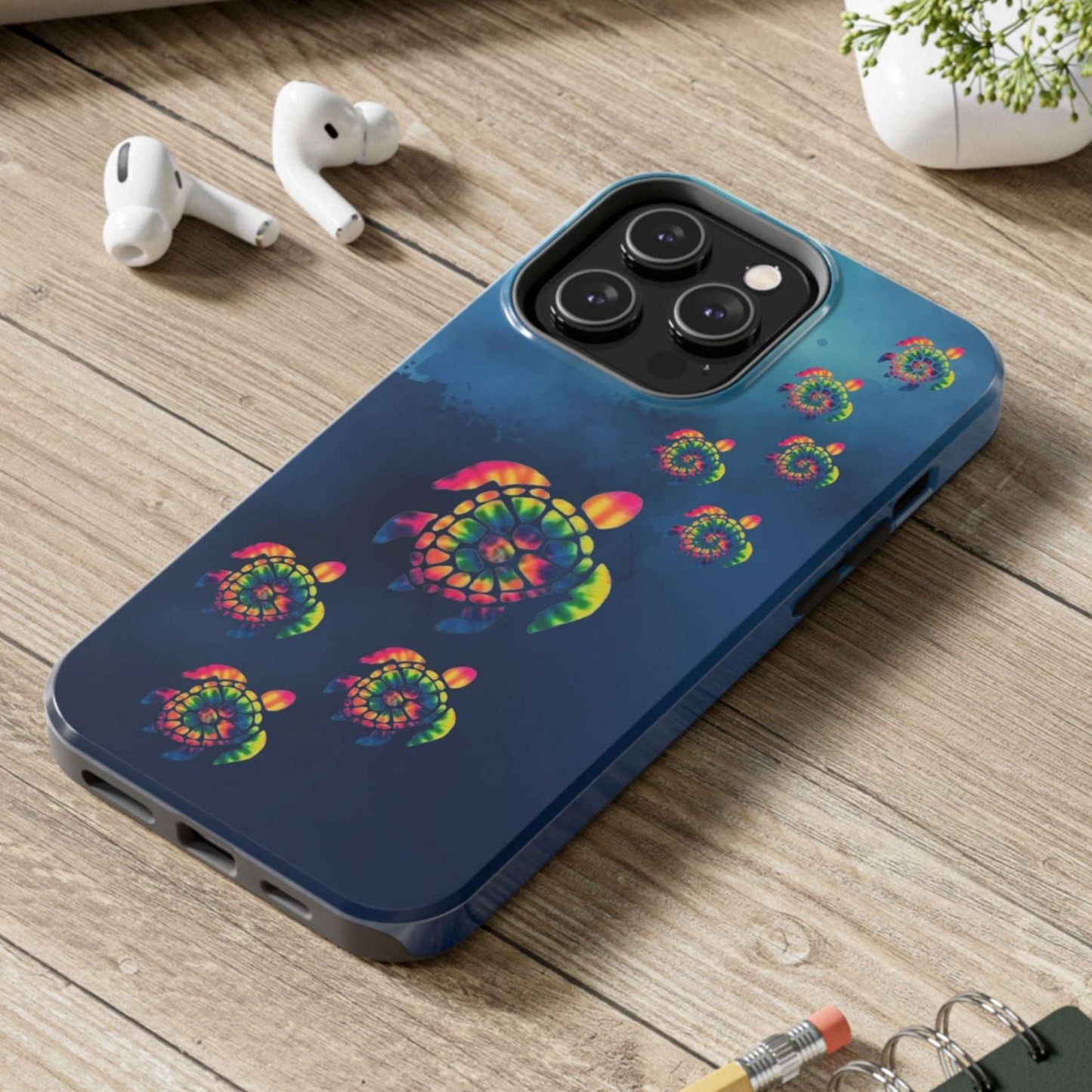 Blue phone case with colorful turtle design on a wooden surface with laptop, earbuds, and notebook.