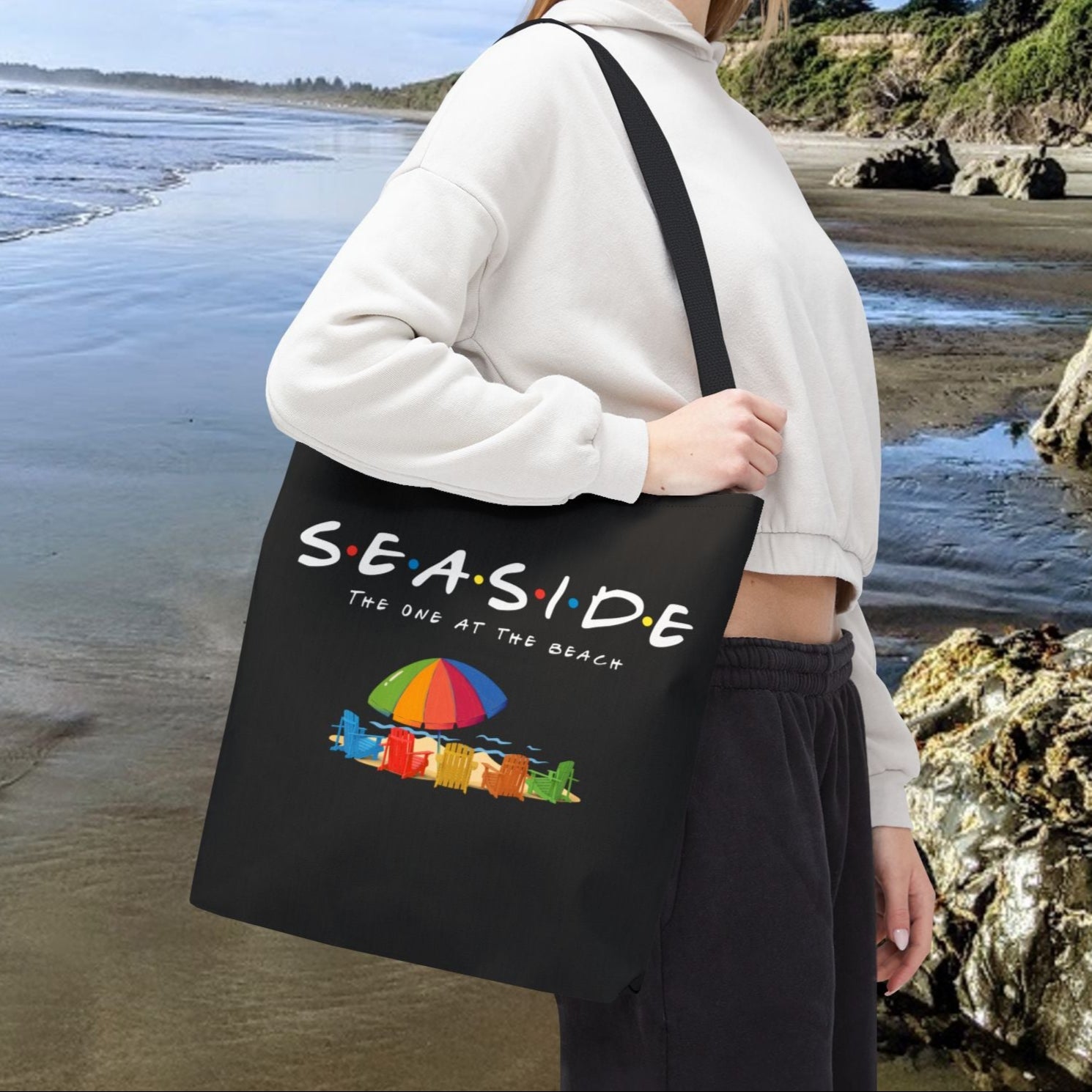 Black 16" x 16" tote bag with the word “Seaside” in rainbow-style letters with colored dots between each letter, underneath says The One at the Beach with five rainbow Adirondack chairs, and a beach umbrella on sand graphic.