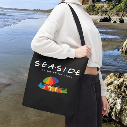 Black 16" x 16" tote bag with the word “Seaside” in rainbow-style letters with colored dots between each letter, underneath says The One at the Beach with five rainbow Adirondack chairs, and a beach umbrella on sand graphic.