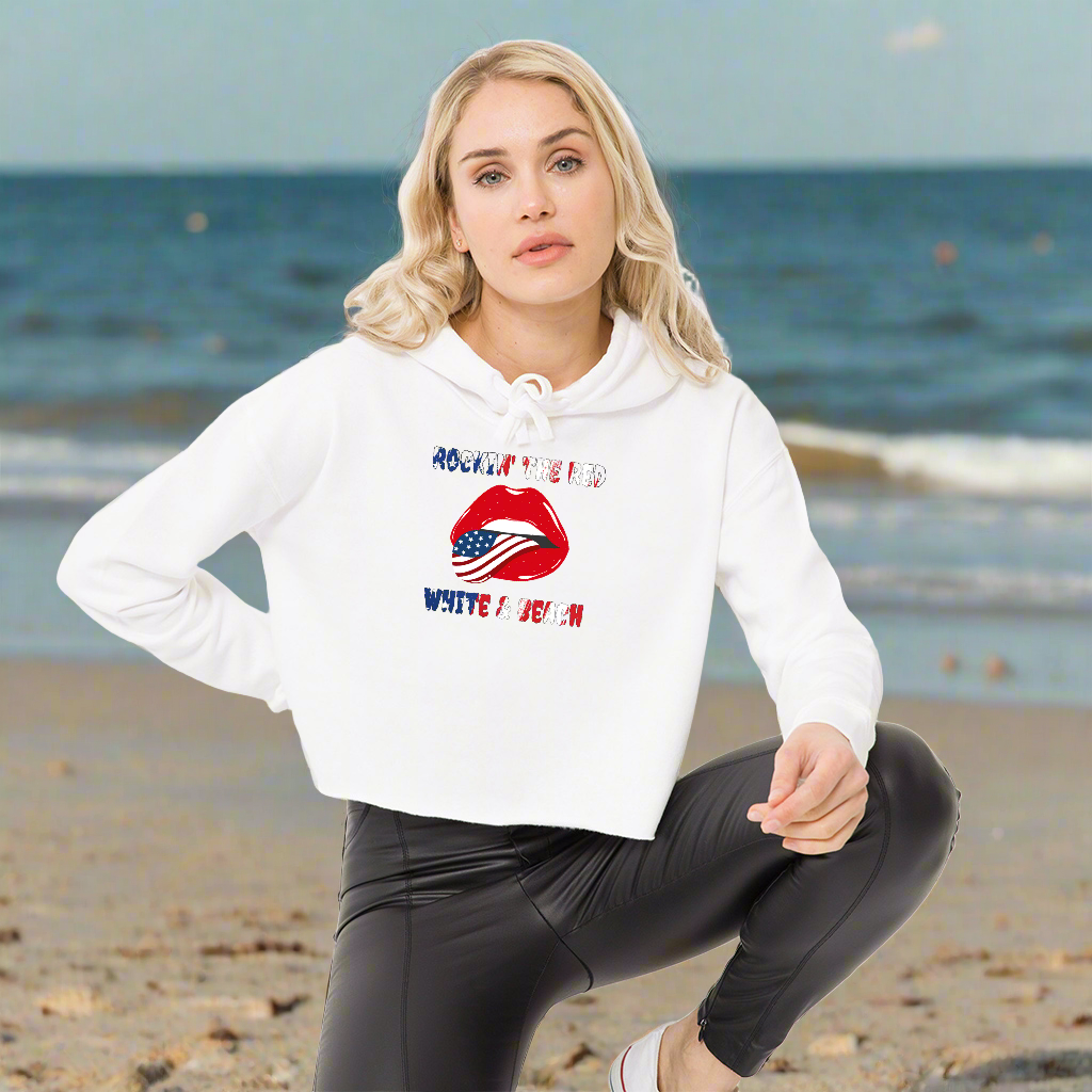 woman kneeling down on the beach in a white cropped hoodie with rolling stone lips with the american flag tongue with flag text that says Rockin' the Red White and Blue 