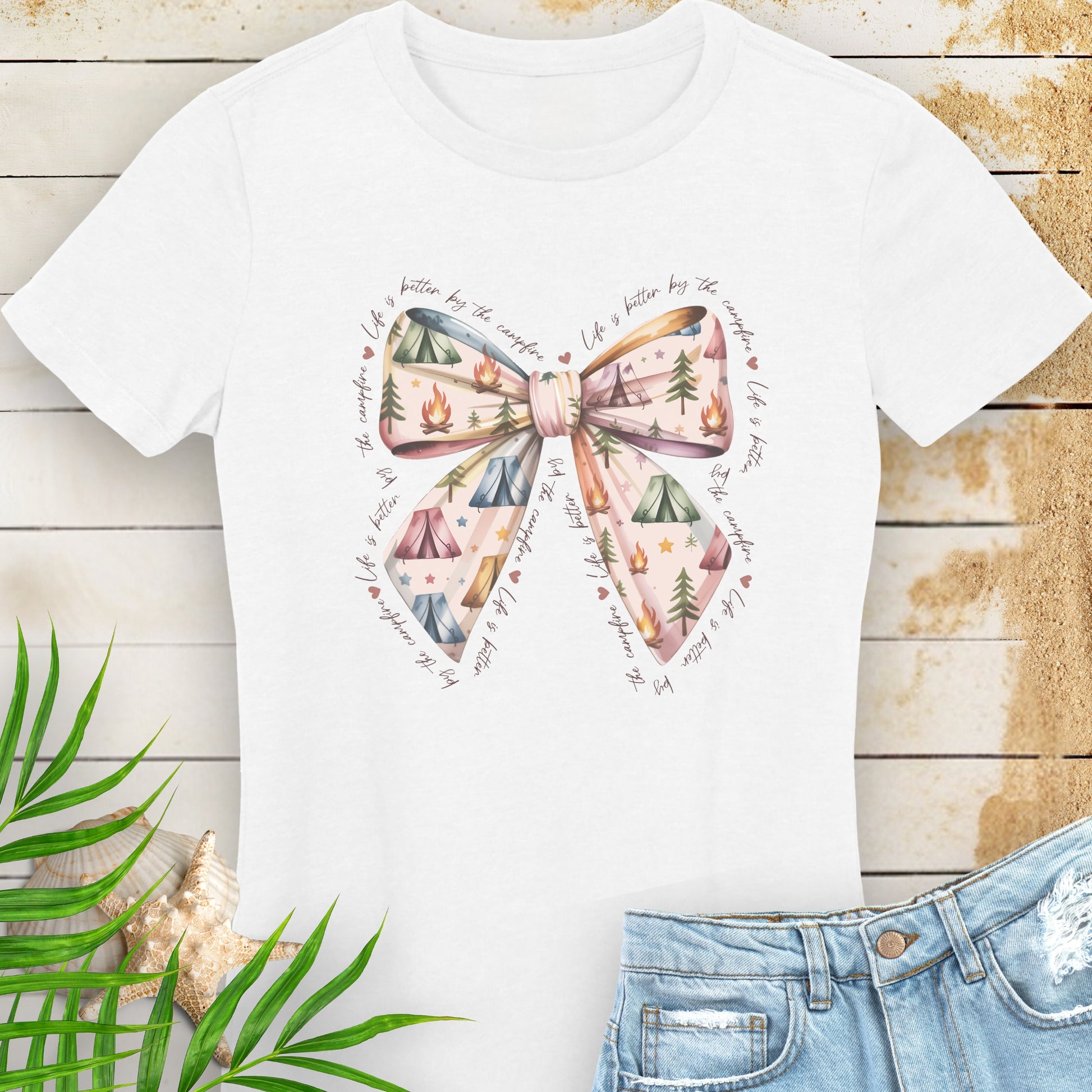 White t-shirt with a bow and nature-themed design on a wooden surface with a leaf and shell.