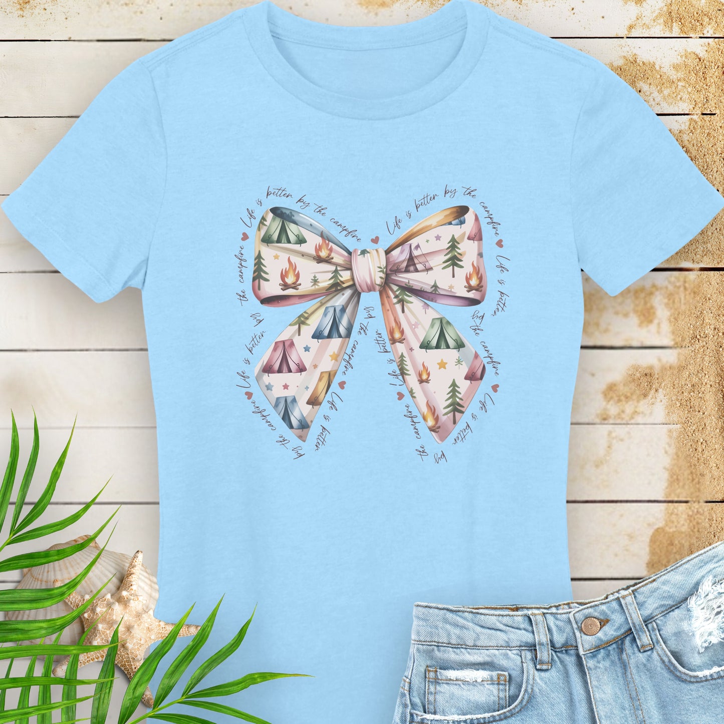 Light blue t-shirt with a bow design on a wooden surface with leaves and shells.