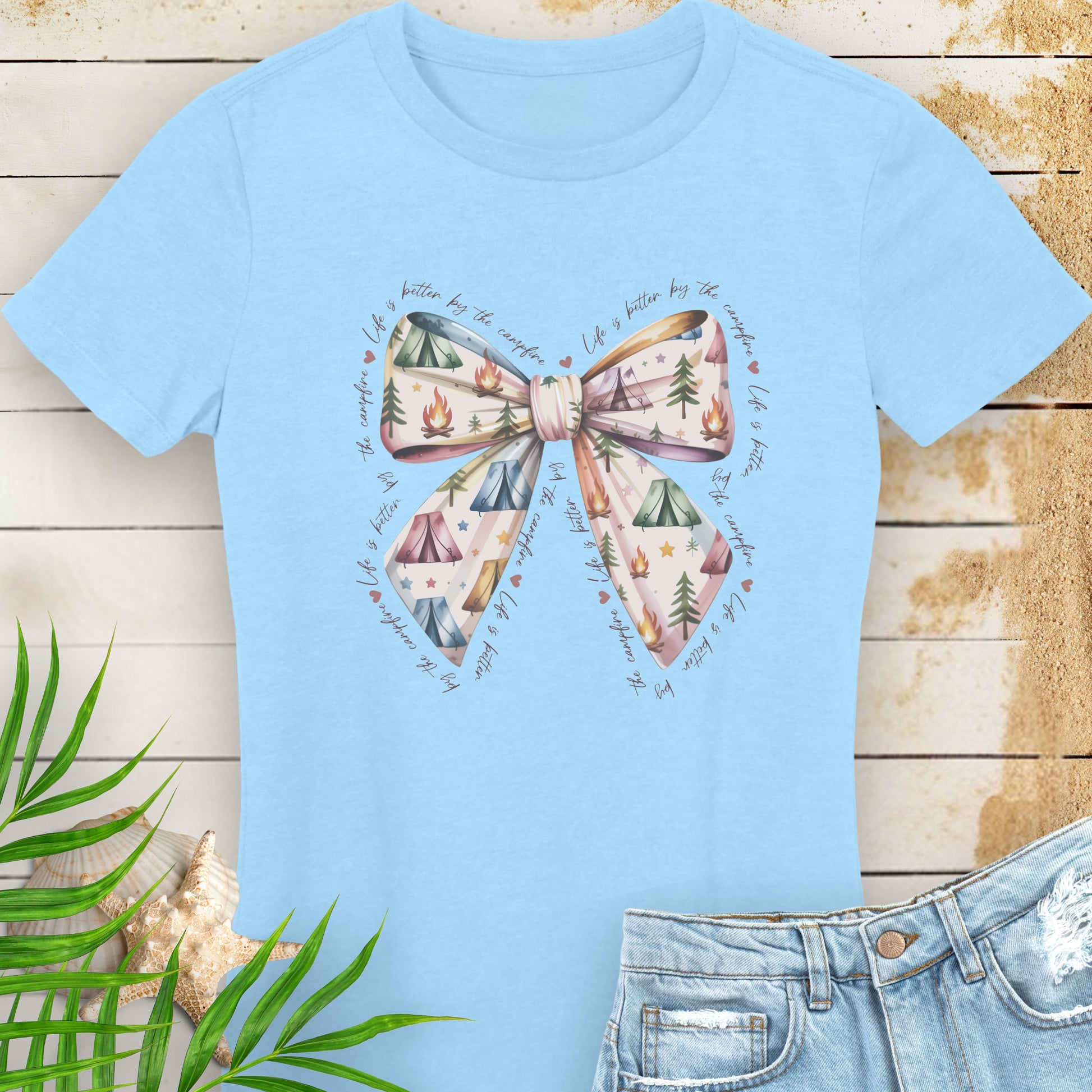 Light blue t-shirt with a bow design on a wooden surface with leaves and shells.