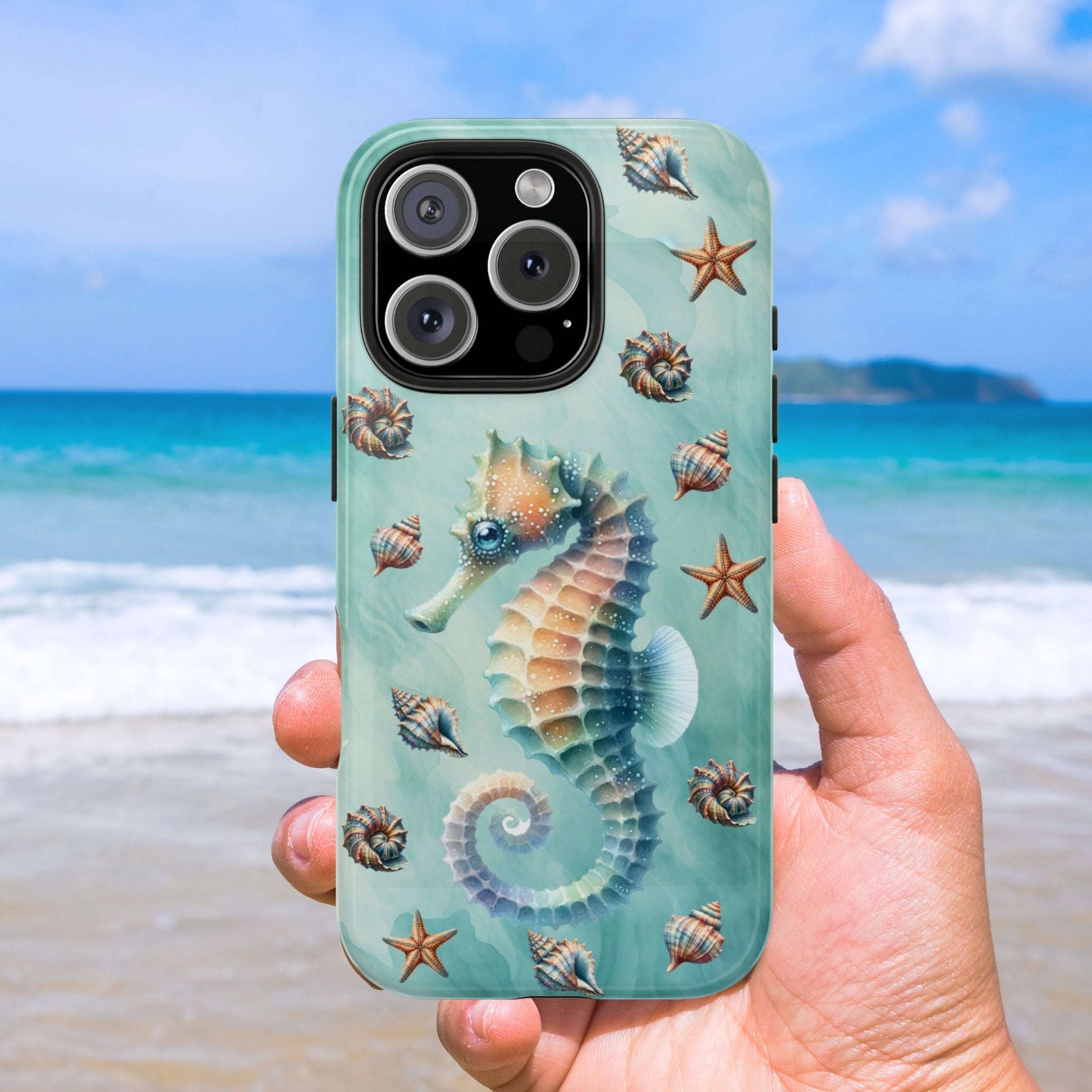 Phone case with seahorse and shell design held by a hand on a beach