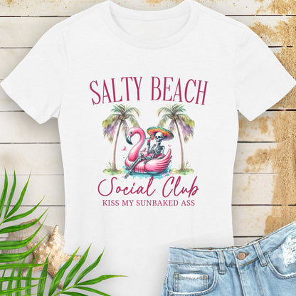 White women’s t-shirt with skeleton in beach outfit floating in a flamingo pool float with palm trees and text “Salty Beach Social Club – Kiss My Sunbaked Ass.”