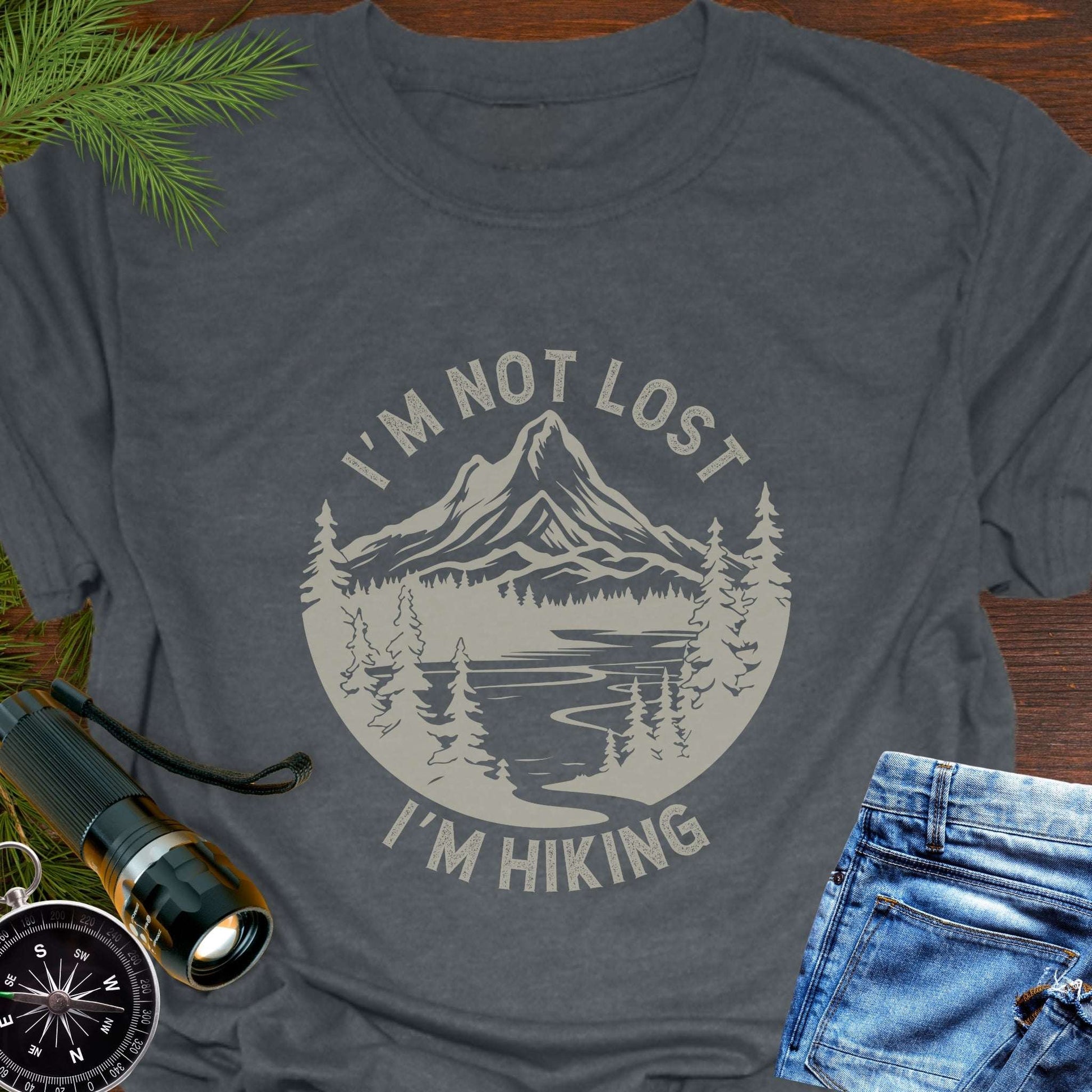 Gray t-shirt with 'I'm not lost, I'm hiking' design on a wooden surface with outdoor accessories.