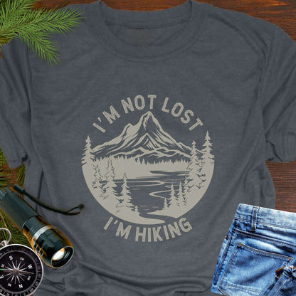Gray t-shirt with 'I'm not lost, I'm hiking' design on a wooden surface with outdoor accessories.
