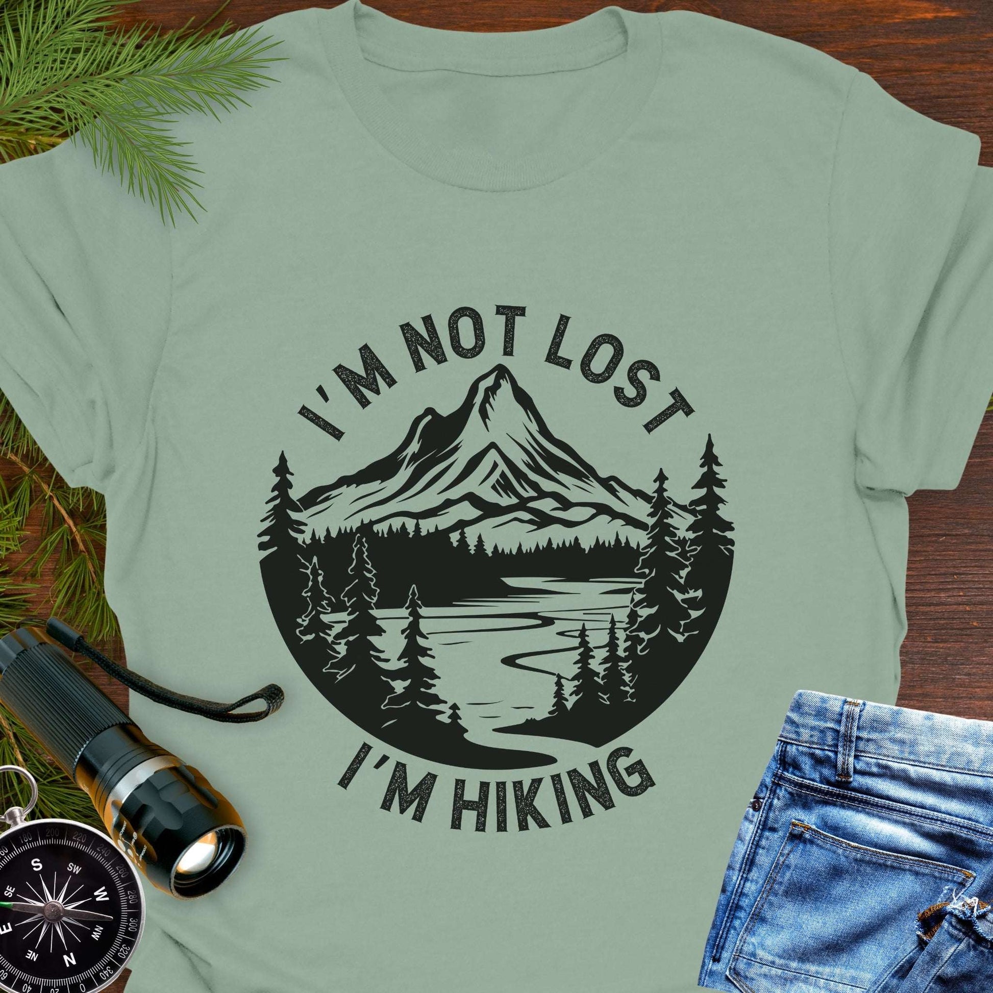 Light green t-shirt with mountain graphic and text 'I'm not lost, I'm hiking' on a wooden surface with outdoor accessories.