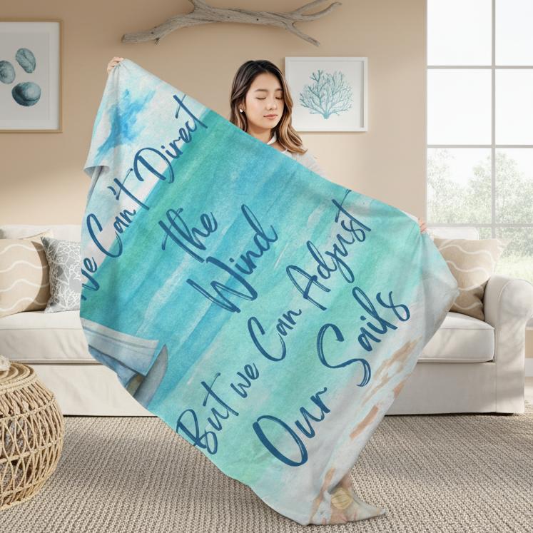 Woman holding Blanket with ocean scene with sailboat sailing from coast with slogan "We Can't Direct the Wind But we Can Adjust Our Sails".