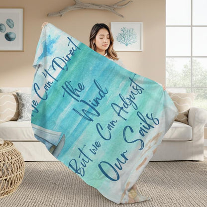 Woman holding Blanket with ocean scene with sailboat sailing from coast with slogan "We Can't Direct the Wind But we Can Adjust Our Sails".
