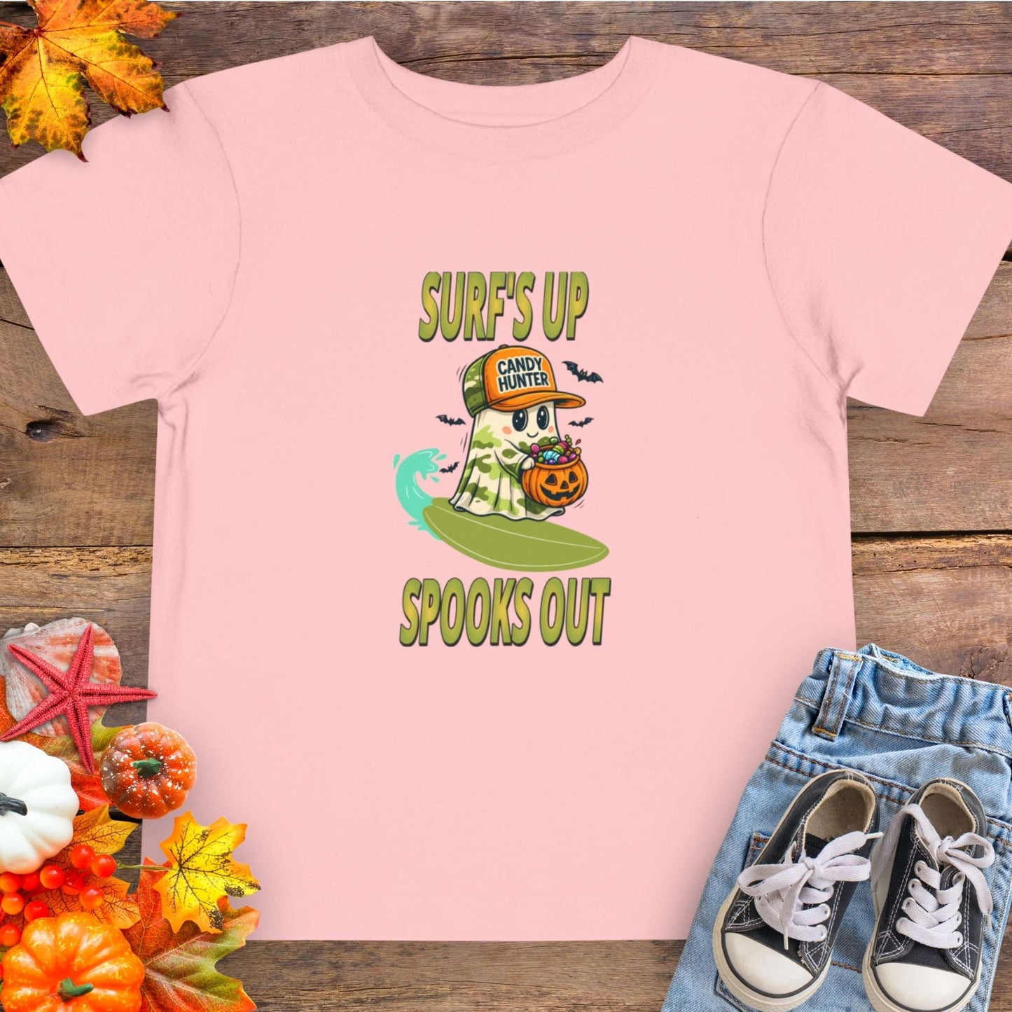 Pink toddler t-shirt with a cartoon ghost child on a surfboard, holding a pumpkin candy bucket, wearing an orange hat labeled “Candy Hunter,” with text that reads “Surf’s Up Spooks Out.”