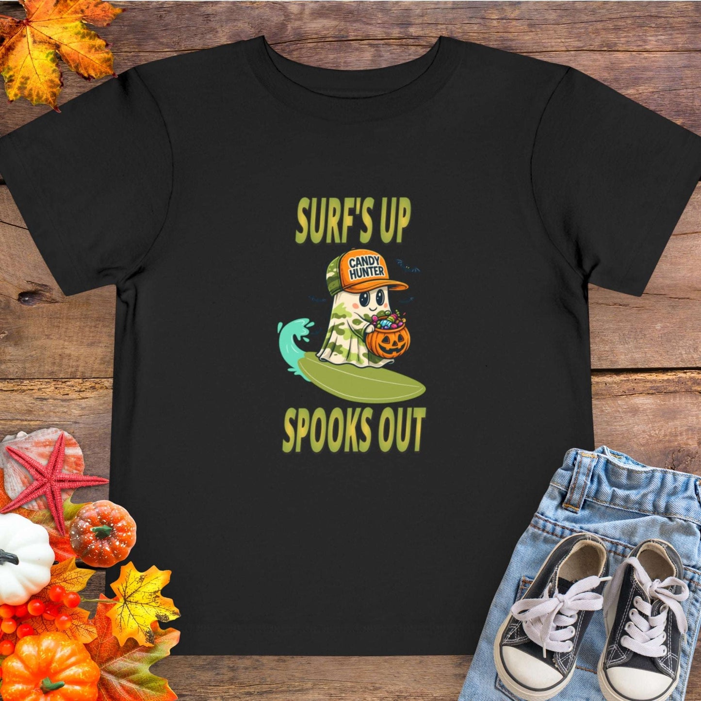 Black toddler t-shirt with a cartoon ghost child on a surfboard, holding a pumpkin candy bucket, wearing an orange hat labeled “Candy Hunter,” with text that reads “Surf’s Up Spooks Out.”