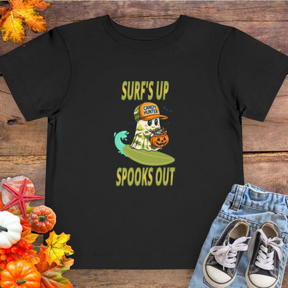 Black toddler t-shirt with a cartoon ghost child on a surfboard, holding a pumpkin candy bucket, wearing an orange hat labeled “Candy Hunter,” with text that reads “Surf’s Up Spooks Out.”