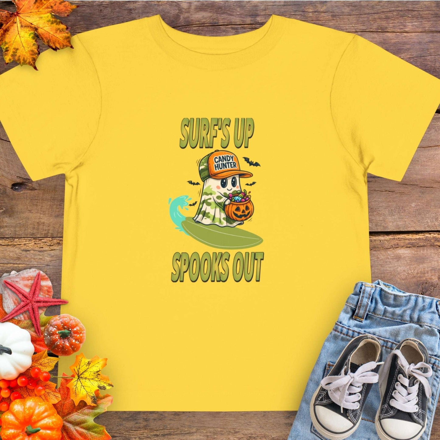 Yellow toddler t-shirt with a cartoon ghost child on a surfboard, holding a pumpkin candy bucket, wearing an orange hat labeled “Candy Hunter,” with text that reads “Surf’s Up Spooks Out.”