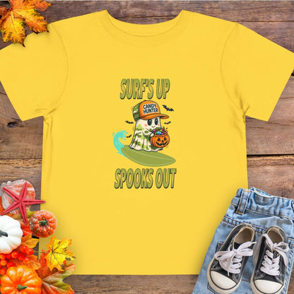 Yellow toddler t-shirt with a cartoon ghost child on a surfboard, holding a pumpkin candy bucket, wearing an orange hat labeled “Candy Hunter,” with text that reads “Surf’s Up Spooks Out.”