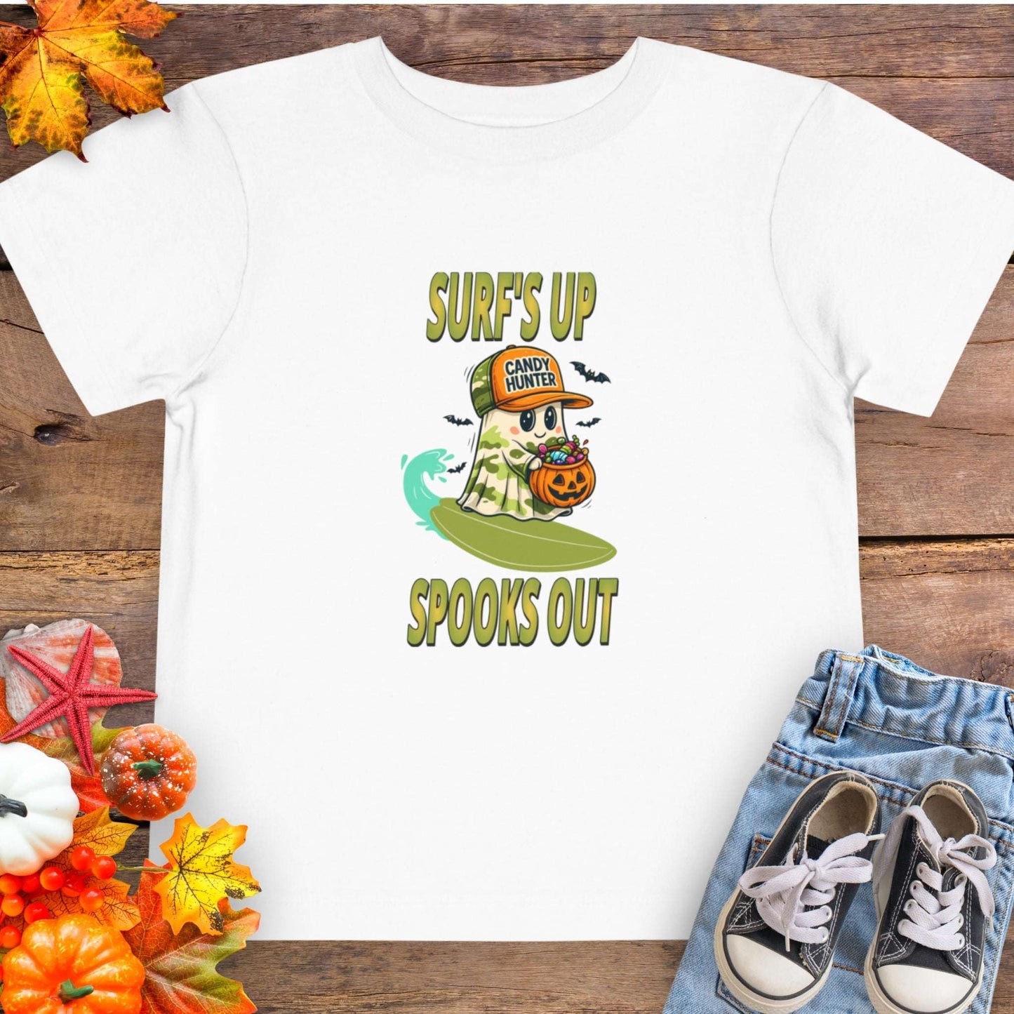 White toddler t-shirt with a cartoon ghost child on a surfboard, holding a pumpkin candy bucket, wearing an orange hat labeled “Candy Hunter,” with text that reads “Surf’s Up Spooks Out.”