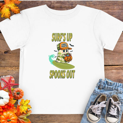 White toddler t-shirt with a cartoon ghost child on a surfboard, holding a pumpkin candy bucket, wearing an orange hat labeled “Candy Hunter,” with text that reads “Surf’s Up Spooks Out.”