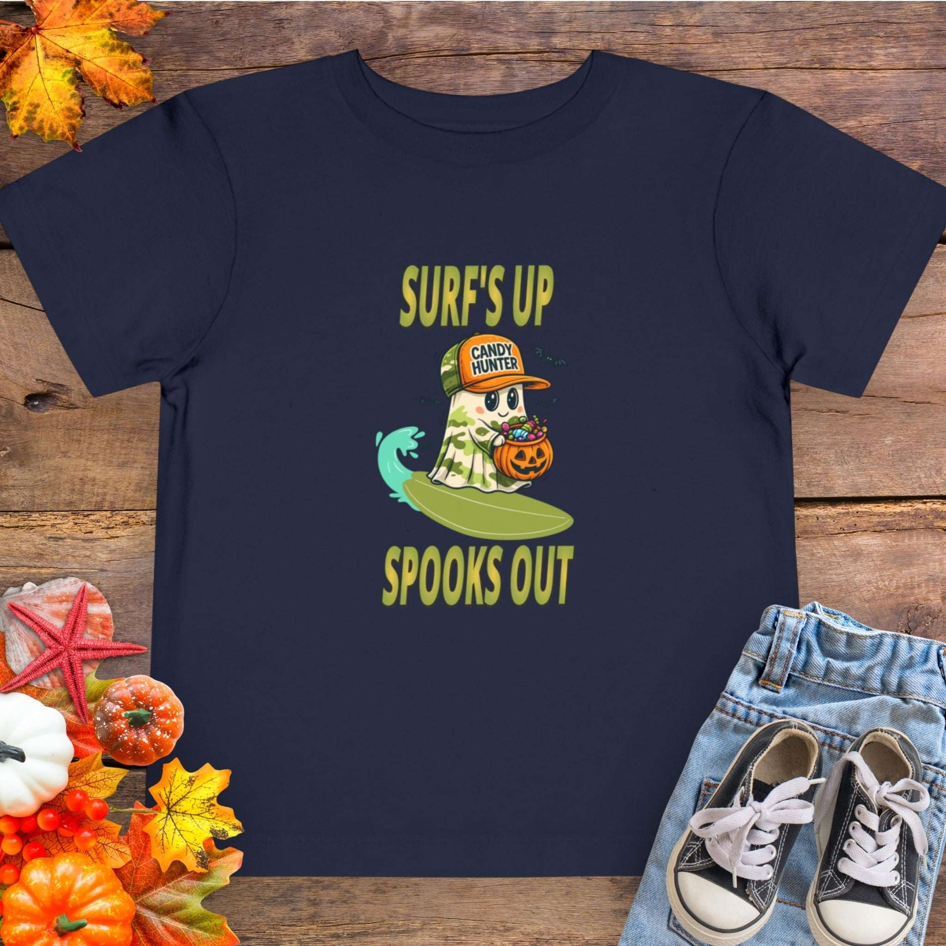 Navy blue toddler t-shirt with a cartoon ghost child on a surfboard, holding a pumpkin candy bucket, wearing an orange hat labeled “Candy Hunter,” with text that reads “Surf’s Up Spooks Out.”