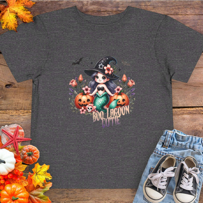 Dark gray Toddler t-shirt design with a witchy mermaid sitting among pumpkins and flowers, with bats flying overhead and the words “Boo Lagoon Cutie.